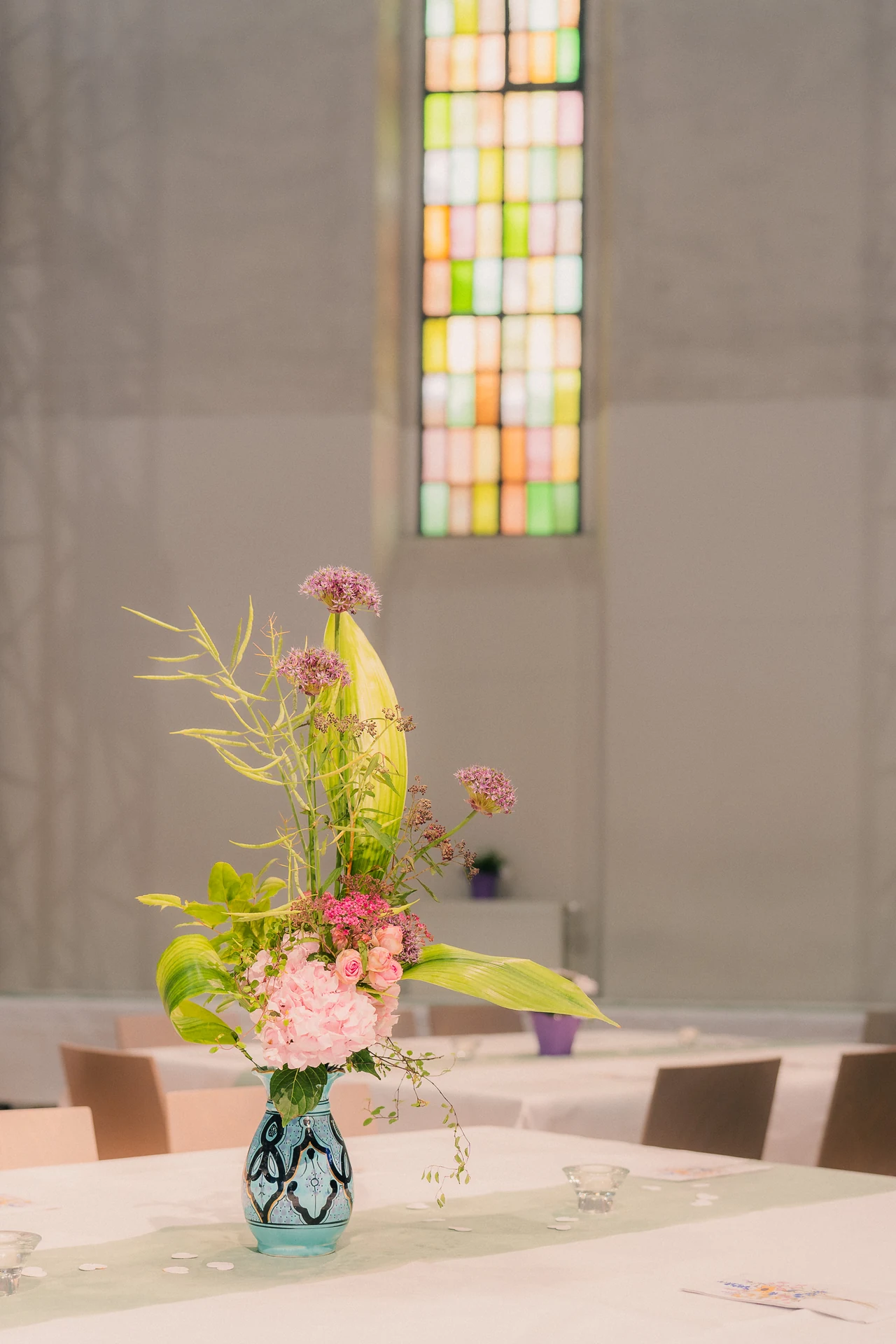 Blumen in einer Glasvase auf dem Tisch, dahinter ein buntes Kirchenfenster