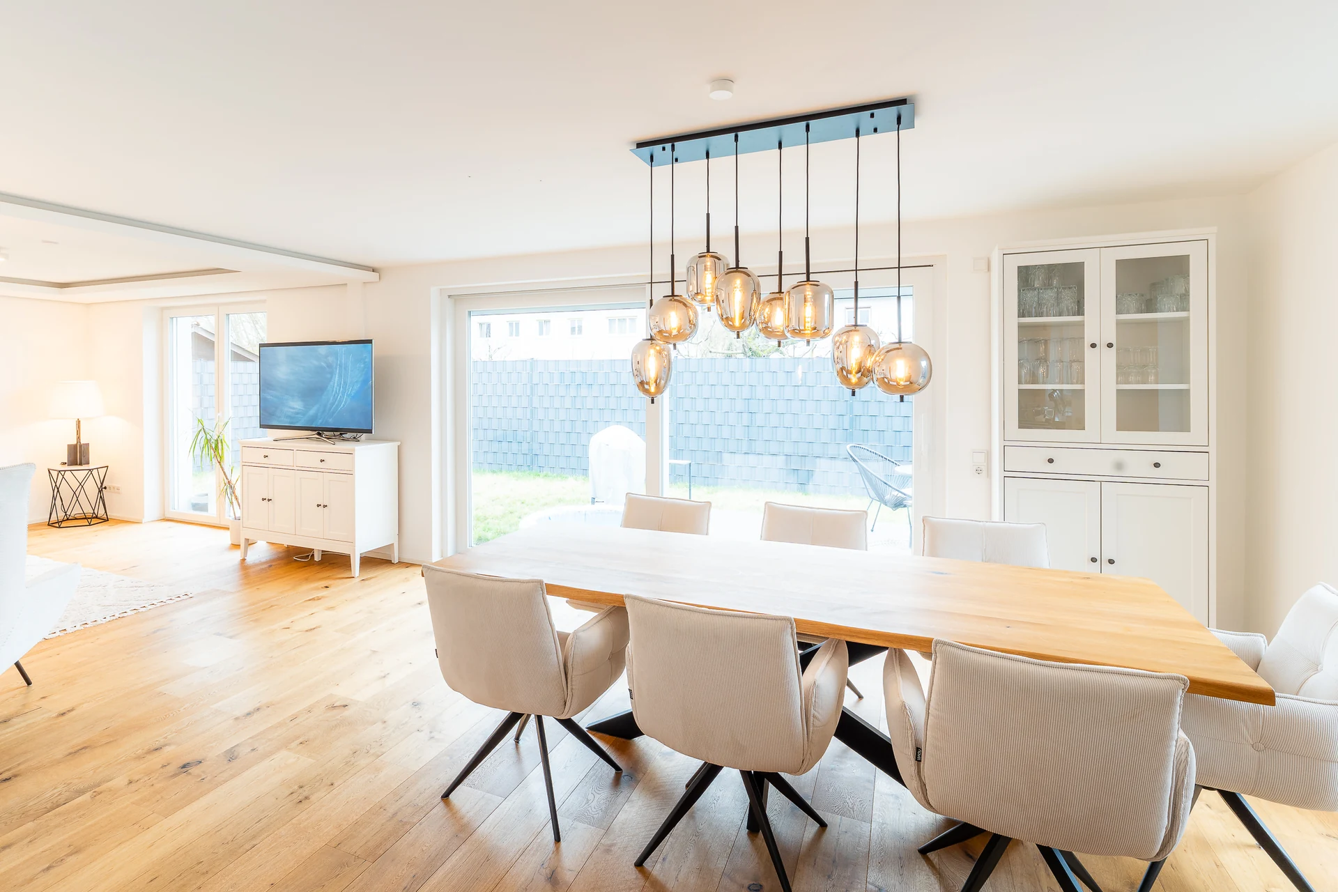 Essbereich mit Holztisch, hellen Stühlen und Pendelleuchte, dahinter das Wohnzimmer