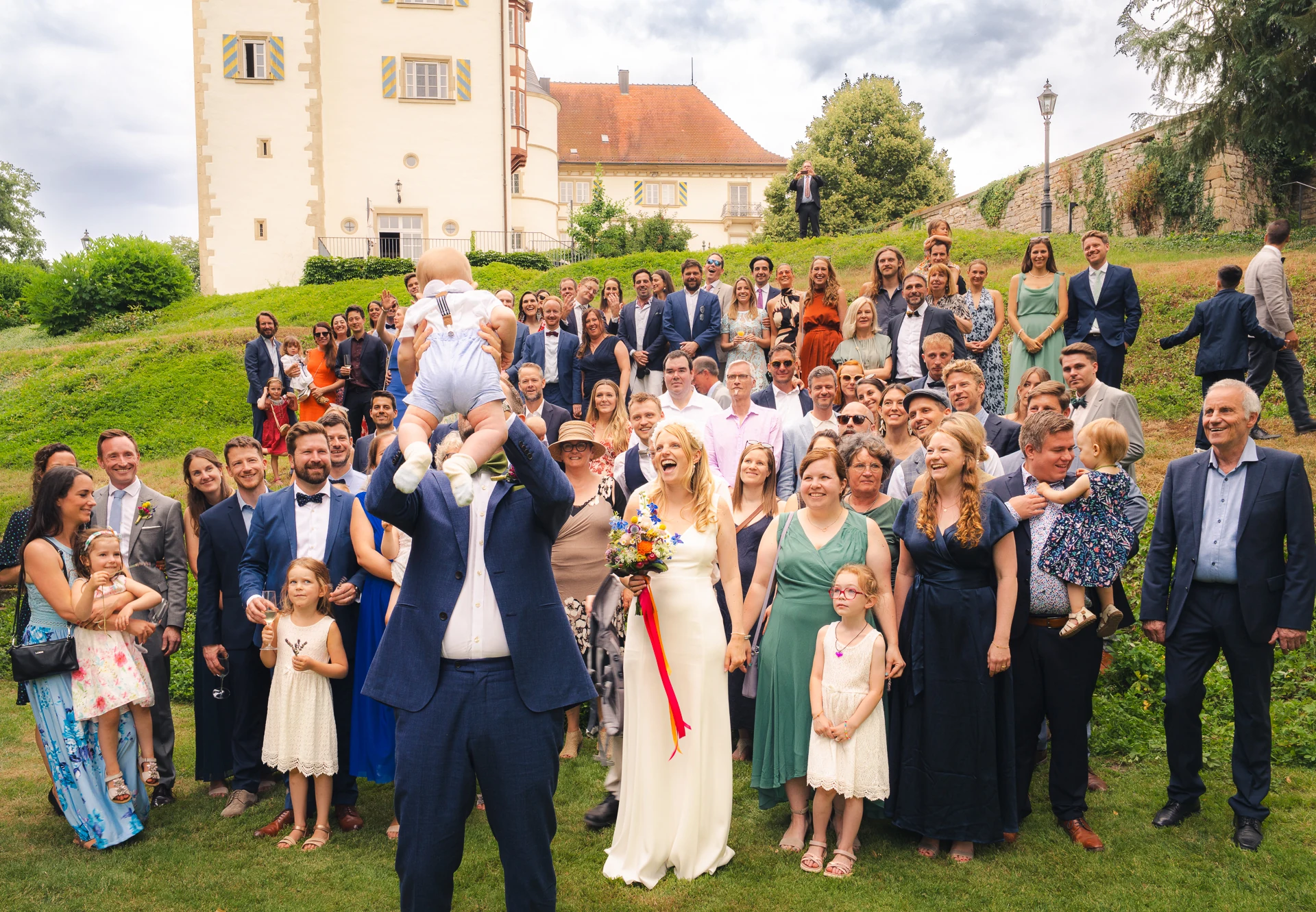 Gruppenbild mit allen Gästen auf der Wiese vor Schloss Neuhaus, ein Kind wird hochgehoben