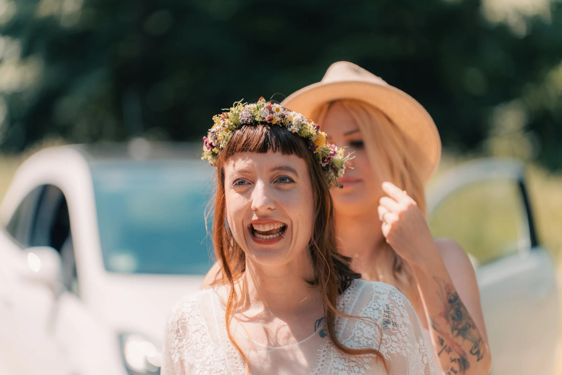 Die Braut mit Blumenkranz lacht, hinter ihr eine Freundin mit Hut