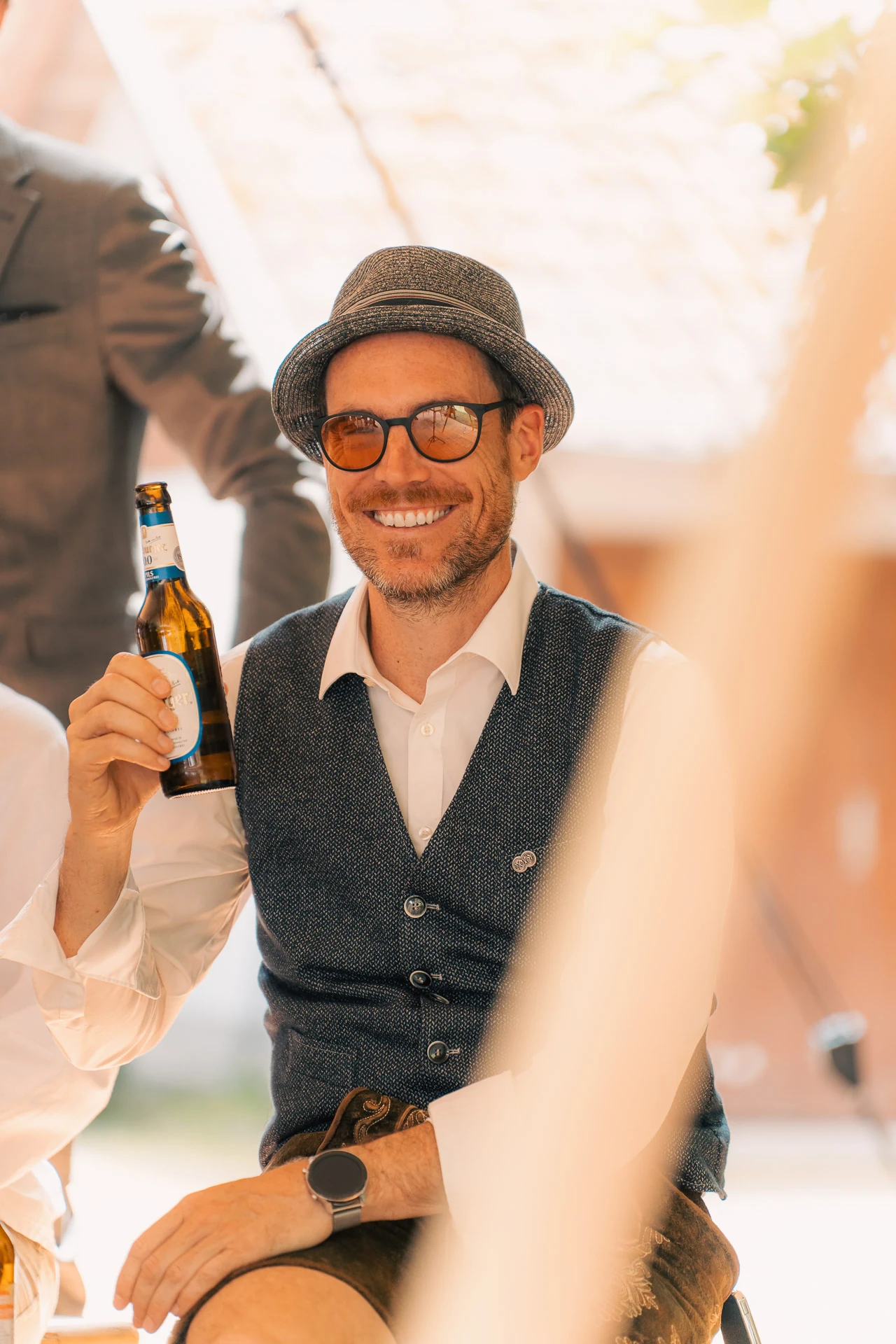 Ein Gast mit Hut und Weste hebt lachend eine Bierflasche