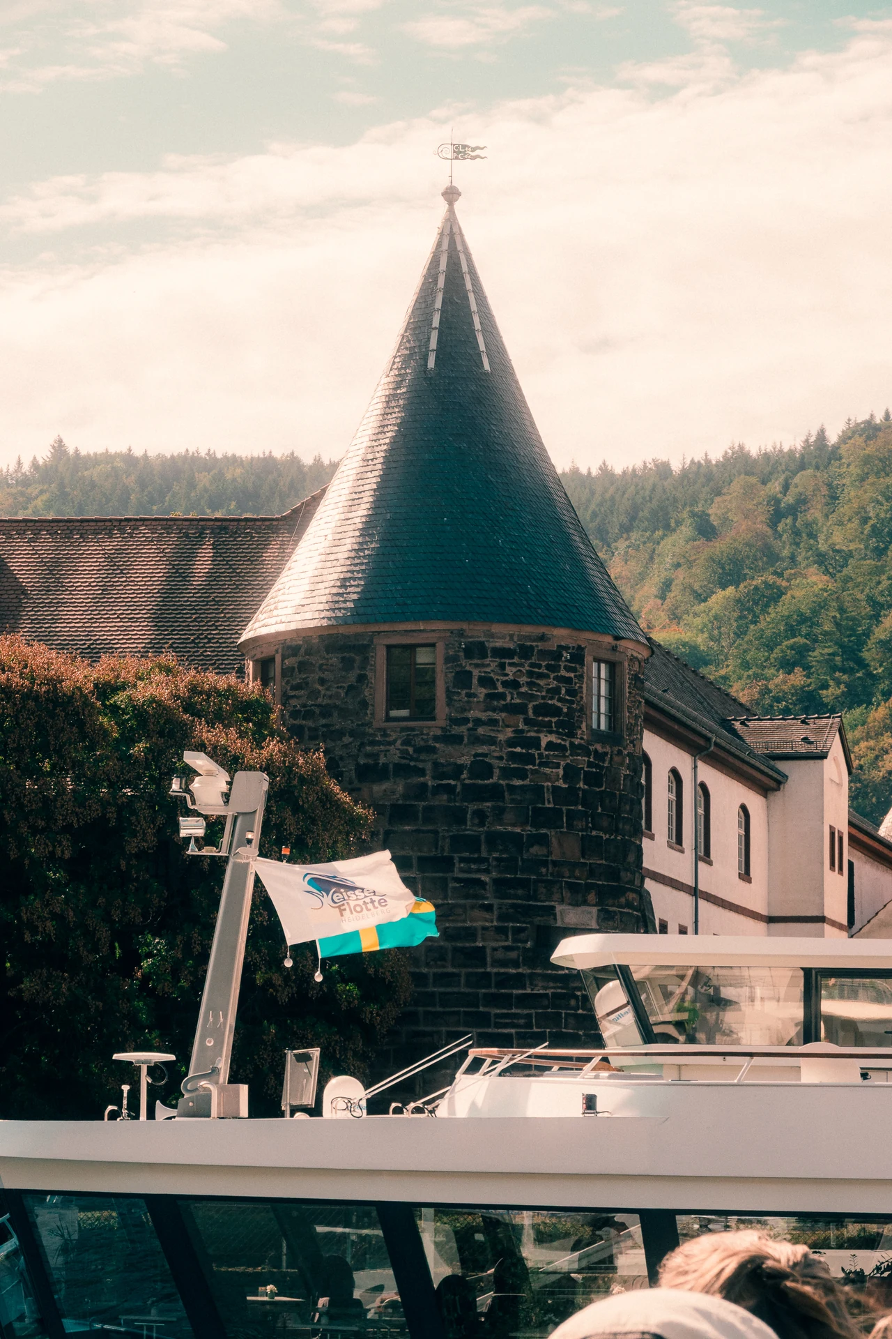 Rundturm mit spitzem Dach am Heidelberger Neckarufer, davor das Oberdeck mit Fahne